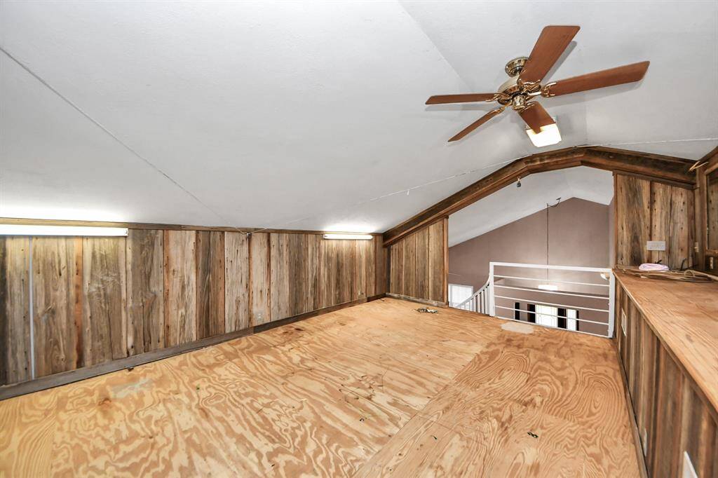 This photo shows a rustic loft space with wood-paneled walls and a slanted ceiling. It features a ceiling fan, ample lighting, and an open railing overlooking the lower level. Ideal for storage or a cozy nook.