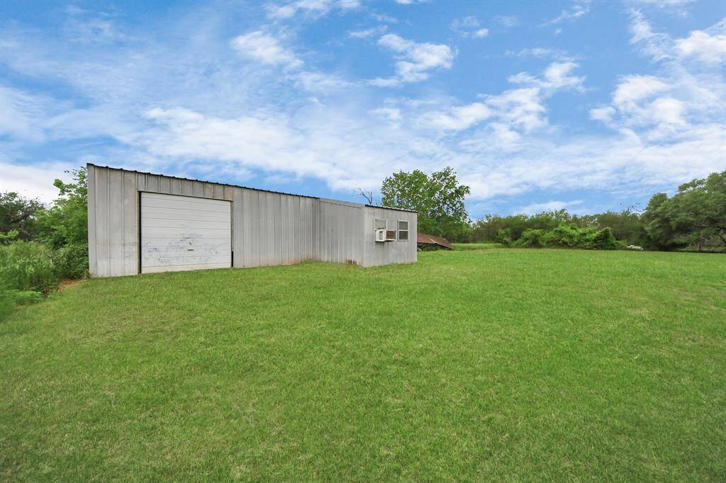 A spacious, grassy yard features a large metal storage building with a roll-up door, perfect for extra storage or a workshop. The area is surrounded by greenery and under a clear blue sky.