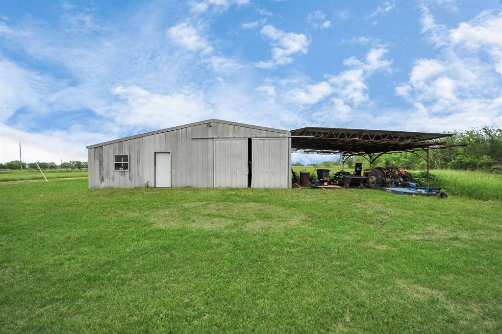 A large metal barn with an attached open shelter sits on a spacious, grassy lot. Ideal for storage or agricultural use, the structure is set against a backdrop of clear skies and open land.