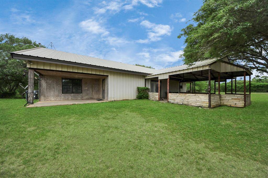 This photo showcases a single-story, metal-roofed home with a rustic exterior, set in a spacious, grassy yard. Two covered patio area with stone accents is visible, providing outdoor living space. The surrounding trees offer a touch of natural privacy.