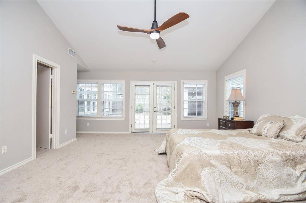 Spacious bedroom with neutral tones, featuring a vaulted ceiling and a modern ceiling fan. Large windows and a glass door provide ample natural light, while plush carpeting adds comfort.