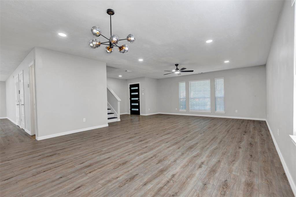 This spacious living area features modern gray flooring, neutral walls, and ample natural light from large windows. It includes a stylish ceiling fixture and a ceiling fan, with an open layout leading to a staircase and hallway.