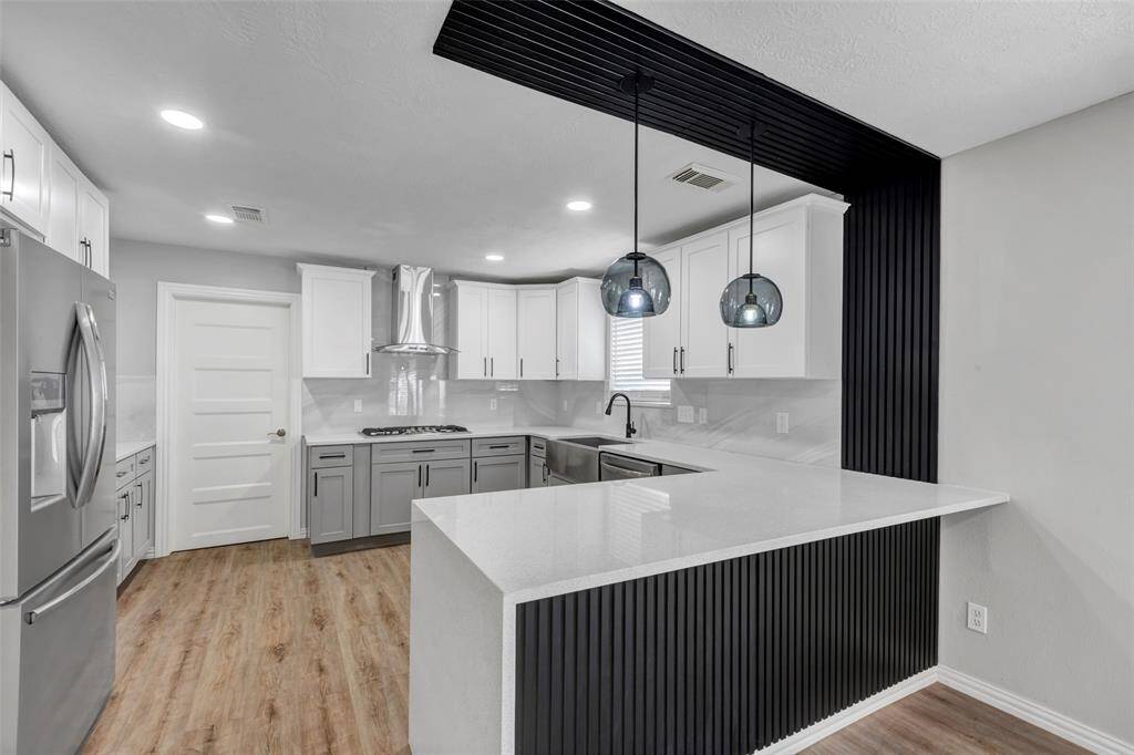 This modern kitchen features sleek white cabinetry, stainless steel appliances, and a spacious island with pendant lighting. It opens to a bright living area with wood-look flooring and large windows, creating a seamless flow for entertaining.