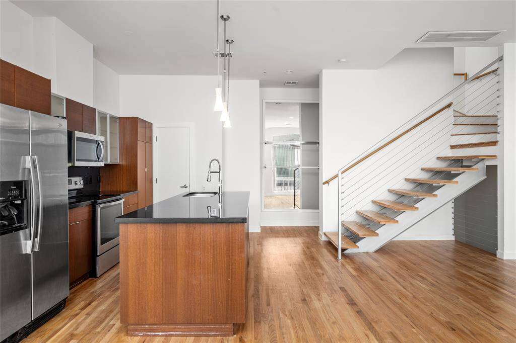 A light hardwood floor covers the space. A wooden staircase with metal railings leads up, its open design showing the support structure.In the center, a modern kitchen island stands with a dark granite countertop, a stainless steel double sink, and a sleek faucet. The island has medium-brown wood cabinets, and a dishwasher is partly visible on the right.