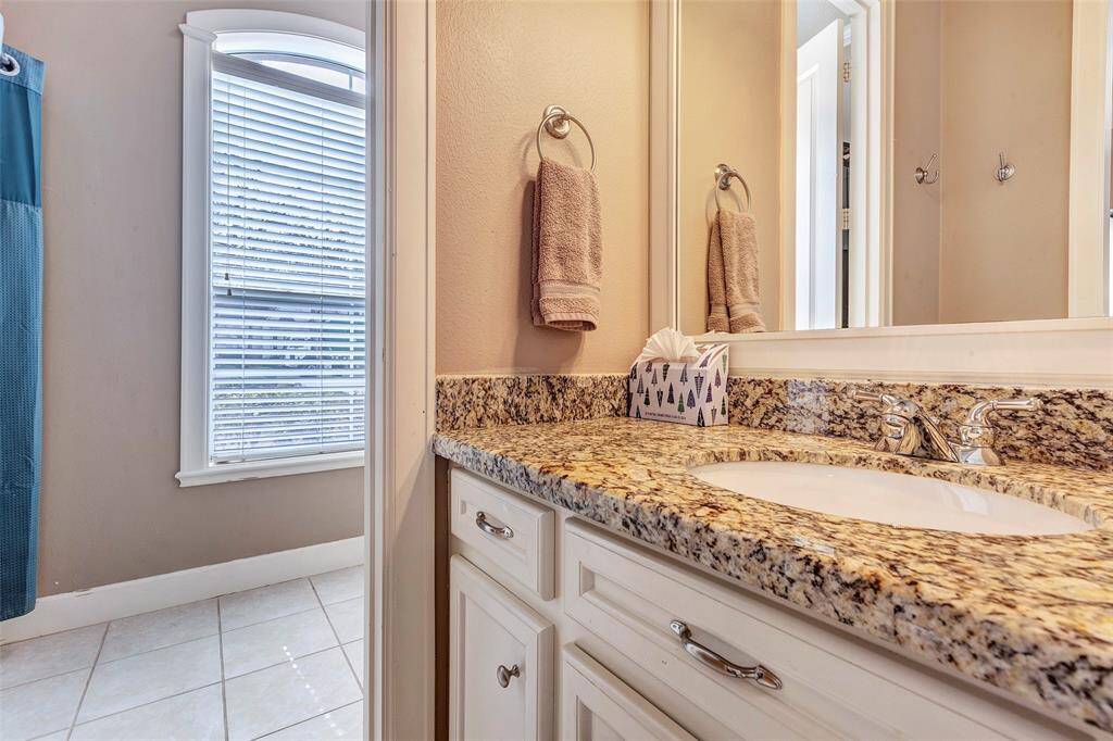 This jack and jill bathroom features a granite countertop with an under-mount sink and elegant fixtures. The space is well-lit by a large window with blinds, and the walls are painted in a neutral tone, complementing the light tile flooring.