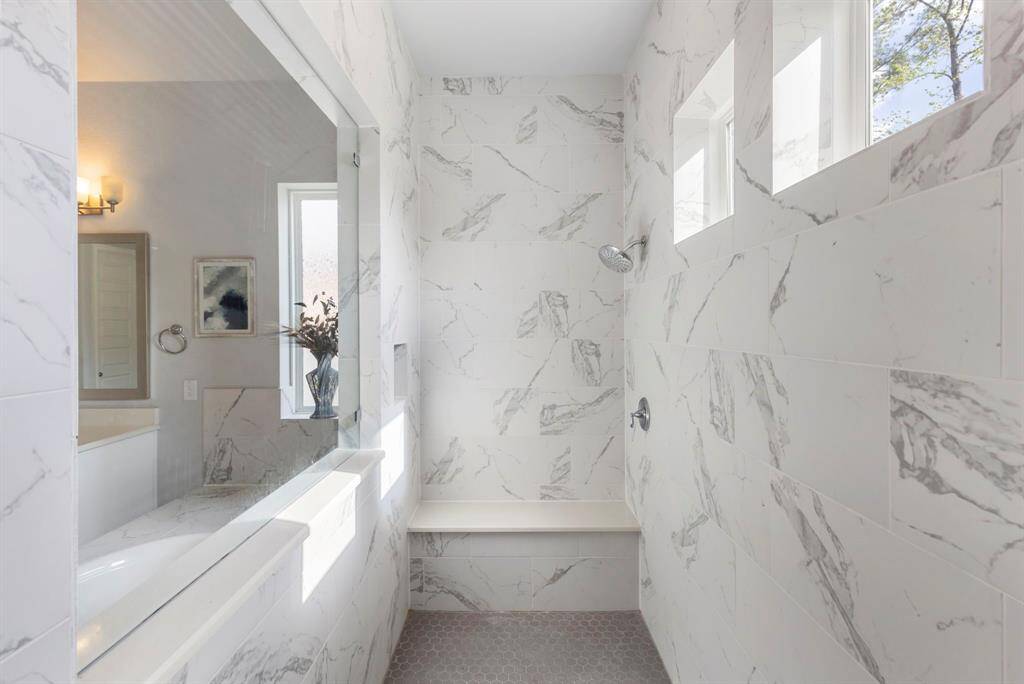 A generous walk-in shower, elegantly wrapped in marbled design tile, is bathed in natural light and offers a serene view of the garden tub.