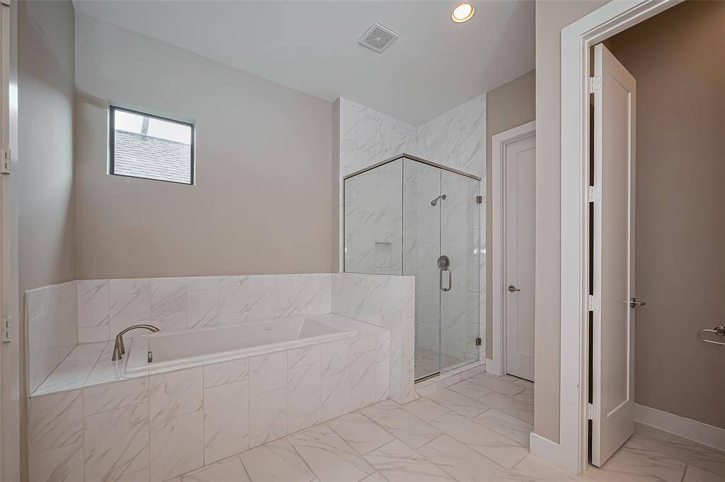 The owner's bathroom boasts a deep, rectangle-shaped massaging air tub, surrounded by quartz tile floors and a matching quartz tub surround. A walk-in glass shower features floor-to-ceiling quartz-tiled walls, with a convenient shampoo niche for added practicality. A window above the tub lets natural light flood the space, while recessed lighting ensures a bright and inviting atmosphere throughout.