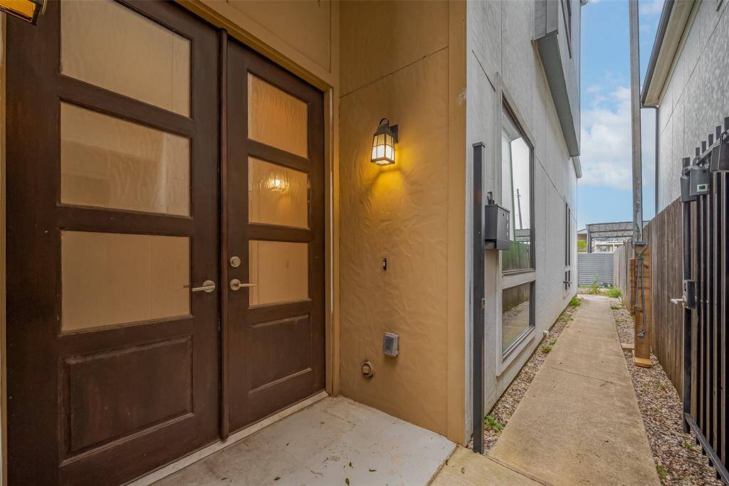 A private walkway leads to the elegant double front doors, featuring modern rainglass inserts that blend style with privacy. Sleek coach lights illuminate the entry, setting a warm, contemporary tone. A wrought iron gate encloses the side and backyard, offering a secure and private outdoor space—perfect for relaxing or entertaining.