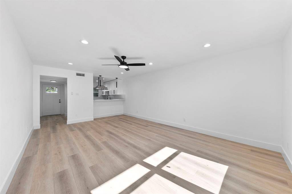 This room features a spacious, open layout with light wood flooring and a neutral color palette. It has recessed lighting, a ceiling fan, and large windows for natural light. The area leads to a modern kitchen with sleek fixtures, creating a bright and inviting space.