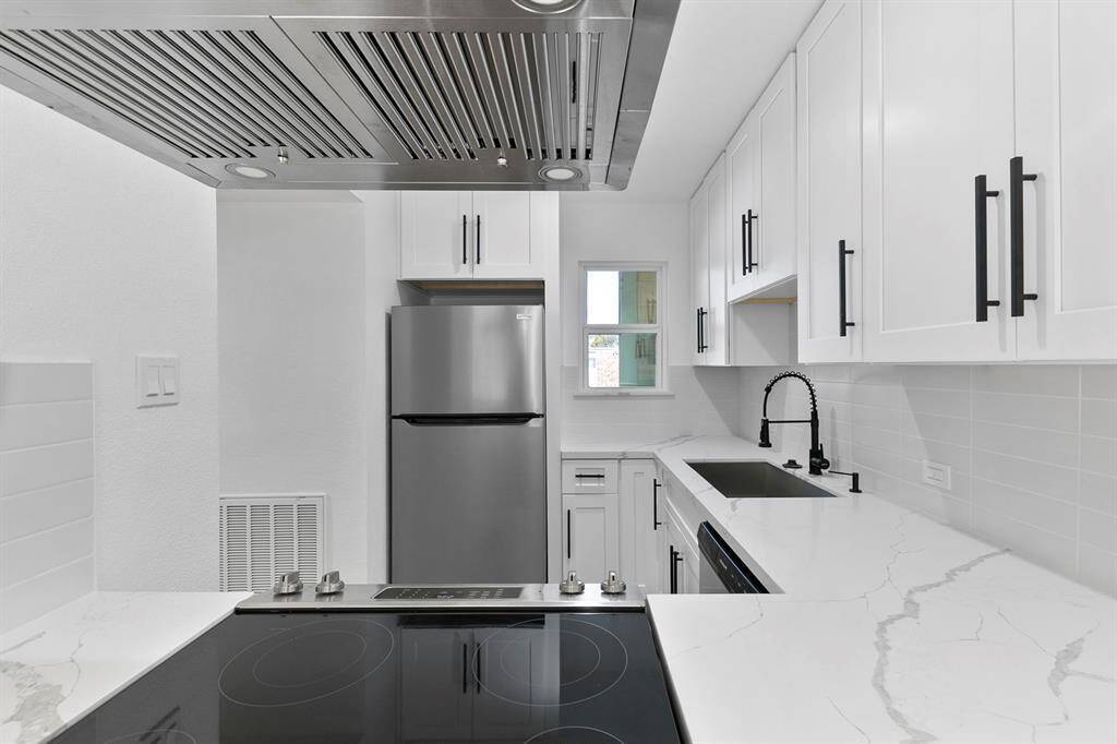 This modern kitchen features sleek white cabinetry with black handles, a stylish black faucet, and a stainless steel refrigerator. It includes an electric cooktop and a minimalist backsplash, offering a clean and contemporary look.