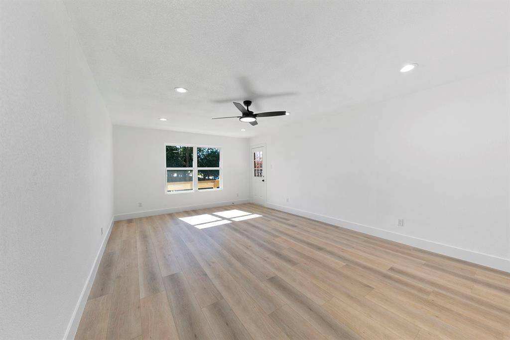 This photo shows a spacious, bright room with light wood flooring and white walls. It features recessed lighting, a ceiling fan, and large windows that let in plenty of natural light.