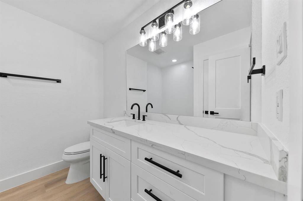 This bathroom features a modern design with a sleek white vanity, marble countertop, and black hardware. It has a large mirror with stylish lighting above, complemented by light wood flooring and a minimalist aesthetic.