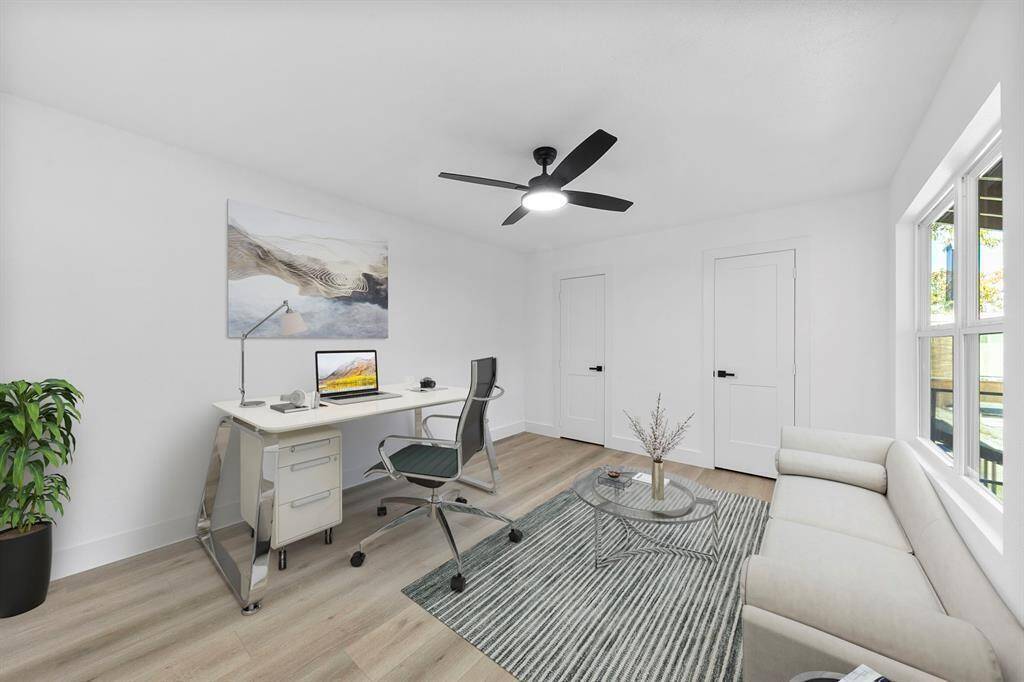 This bright, modern home office features sleek furnishings, wood flooring, and ample natural light from large windows. The room includes a stylish desk, ergonomic chair, cozy sofa, and a ceiling fan for comfort.