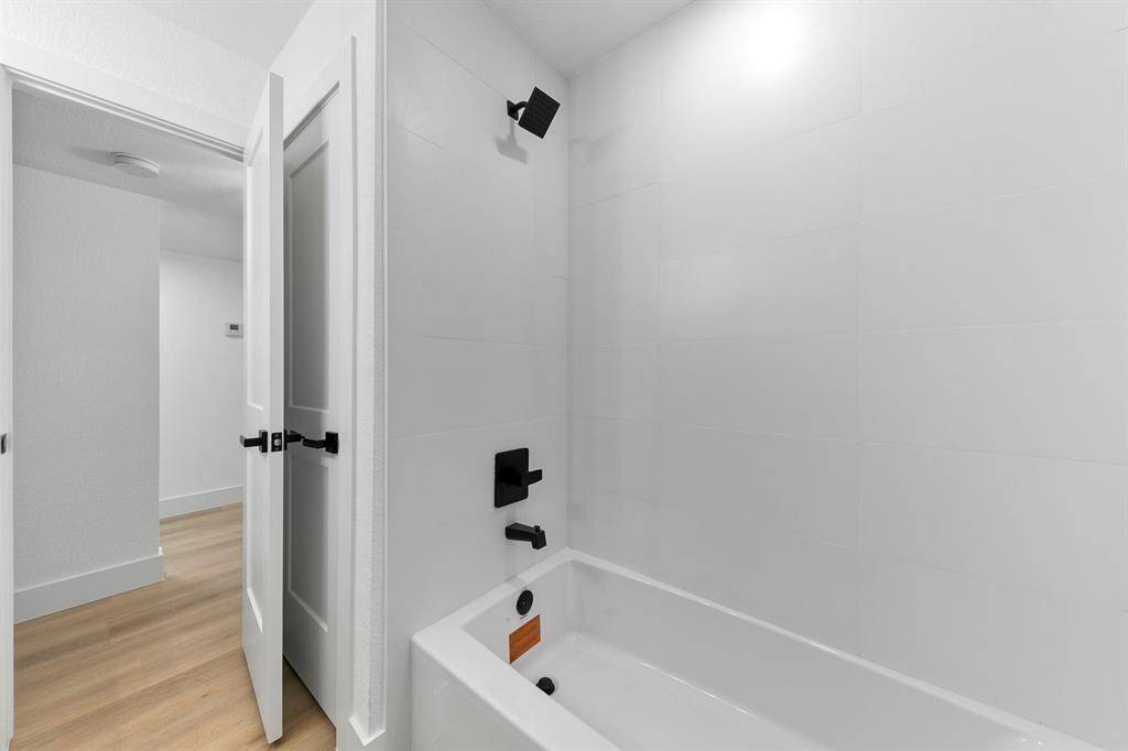 This photo showcases a modern bathroom with a clean, minimalist design. It features a white tiled bathtub with sleek black fixtures and a partially open door leading to a room with light wood flooring.