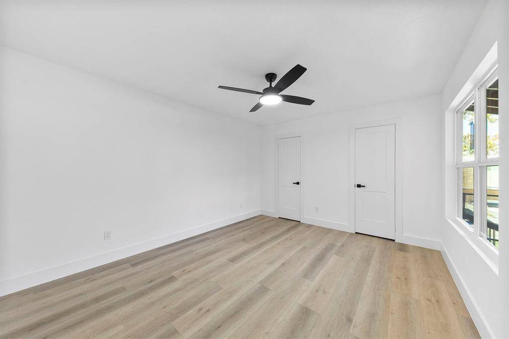 This is a bright, minimalist room with white walls and light wood flooring. It features a modern black ceiling fan, two white doors, and a window allowing natural light to fill the space.