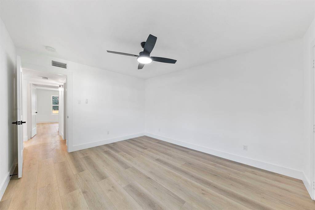 This photo shows a bright, empty room with light wood flooring and white walls. It features a modern ceiling fan and is connected to a hallway, promoting an open and airy feel.