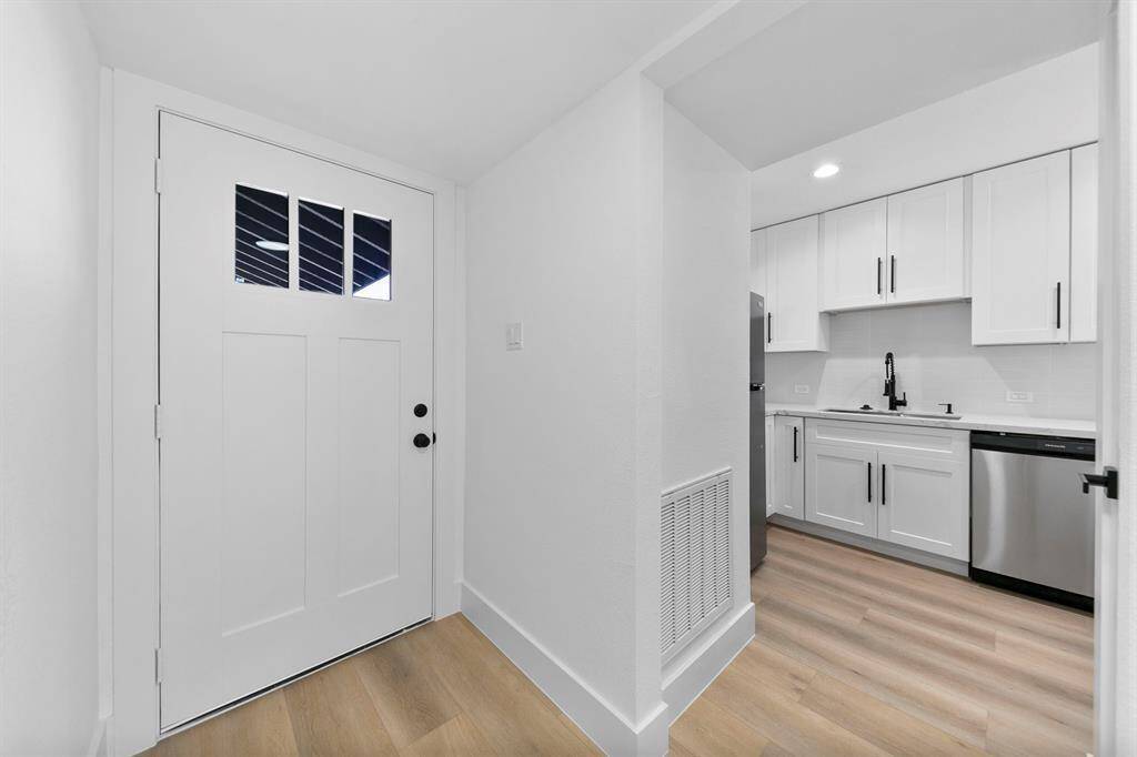 This photo shows a modern, bright entryway leading to a sleek kitchen. The space features a white door with windows, light wood flooring, and a kitchen with white cabinetry, stainless steel appliances, and black hardware.