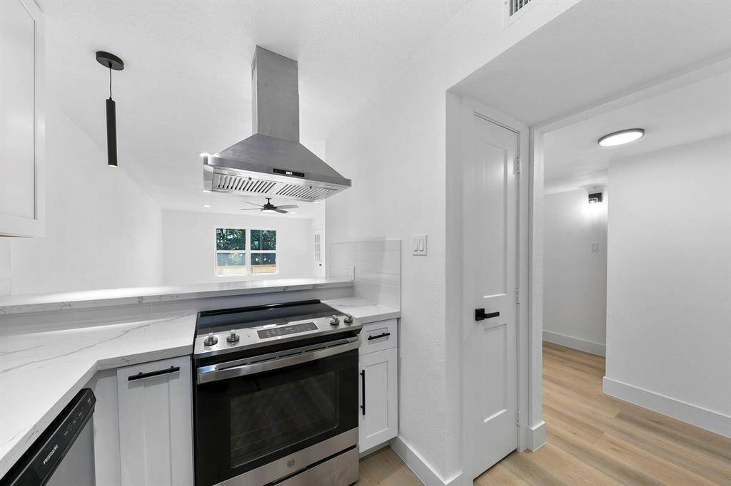 This kitchen features a modern, sleek design with white cabinetry, stainless steel appliances, and a central island hood. It has light wood flooring and ample natural light from the window, creating a bright and airy feel.