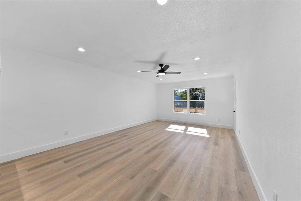 Bright, spacious room with light wood flooring, white walls, and a ceiling fan. Large window offers natural light. Ideal for living or family room.