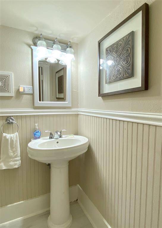 This is a compact, well-lit bathroom featuring a pedestal sink, a wall-mounted mirror with overhead lighting, and decorative wainscoting. It has a clean, classic design with neutral tones and tasteful wall art.