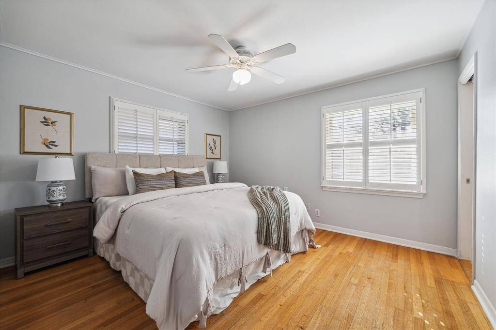 The relaxing primary bedroom is physically staged here with a king sized bed. The room displays a large window to the backyard, original hardwood floors, wood plantation-style shutters and a full bathroom.