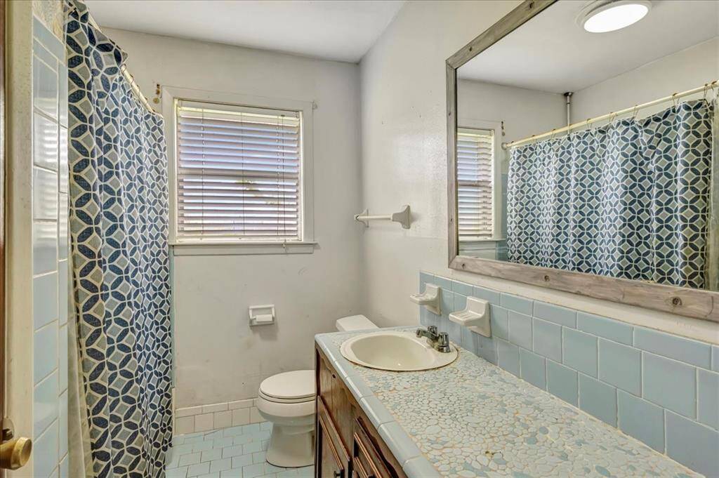 This bathroom features vintage blue tile, a large mirror, and a mosaic countertop. It has a bathtub with a patterned shower curtain and a window for natural light.