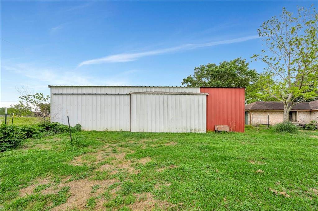 The photo shows a large metal barn with white and red siding, set in a spacious grassy yard. The property features open skies and mature trees, providing a rural, peaceful setting.