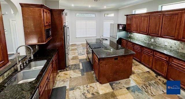 Spacious kitchen with dark granite countertops and rich wooden cabinetry. Features an island with a cooktop, stainless steel appliances, and tiled flooring. Bright with ample natural light from high-set windows.