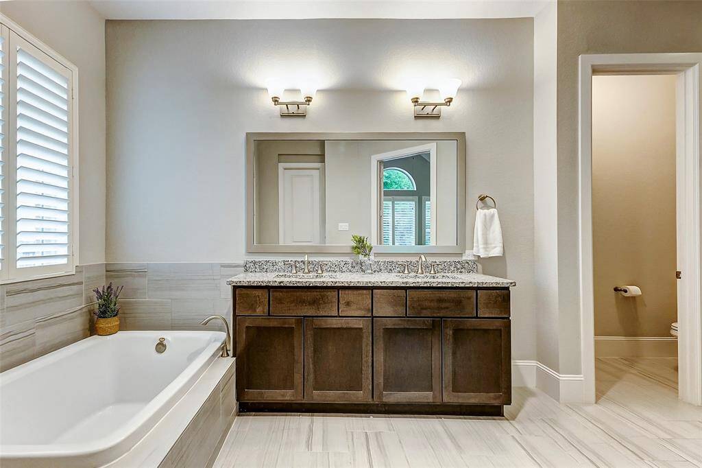 Step through the double doors into your spacious ensuite bathroom, where luxury meets functionality. A large vanity with dual sinks, granite countertops, a framed mirror, and striking lighting fixtures set the tone for elegance. To the left, the soaking tub is perfectly positioned beneath a large window with plantation shutters, offering a tranquil spot to relax and unwind.
