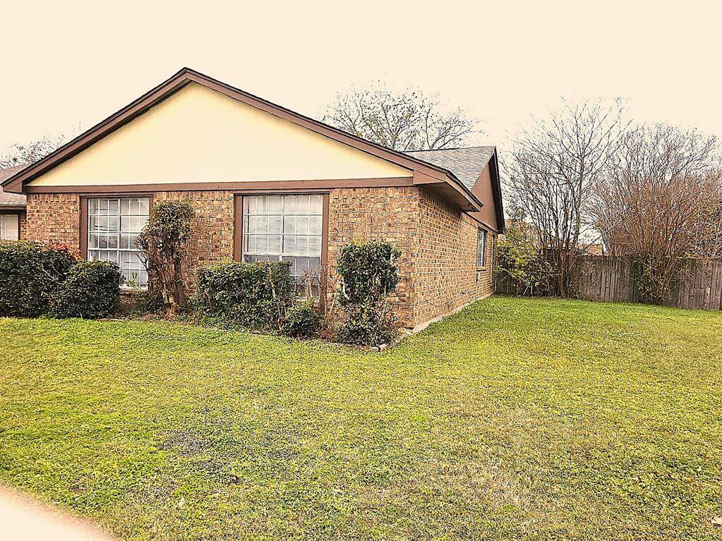 Single-story house with a sloped roof. It features large front windows and is surrounded by a well-maintained lawn and trimmed shrubs, with a wooden fence enclosing the backyard.