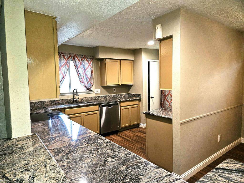 This kitchen features a modern design with granite countertops and light wood cabinets. It includes stainless steel stove and disher washer, a sleek sink, and natural light from a window with patterned curtains. The space has a warm, inviting feel with tile flooring.