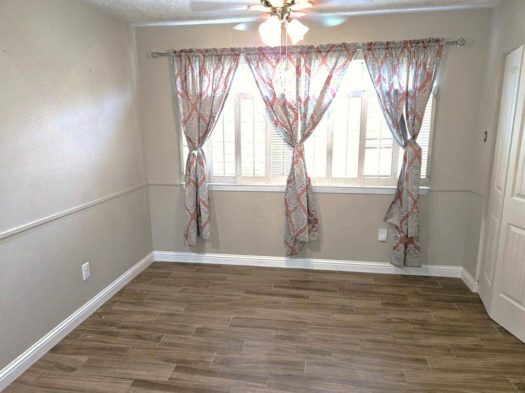 This room features tile-wood style flooring, neutral walls, and a window with floral curtains allowing natural light. A ceiling fan is present for comfort.