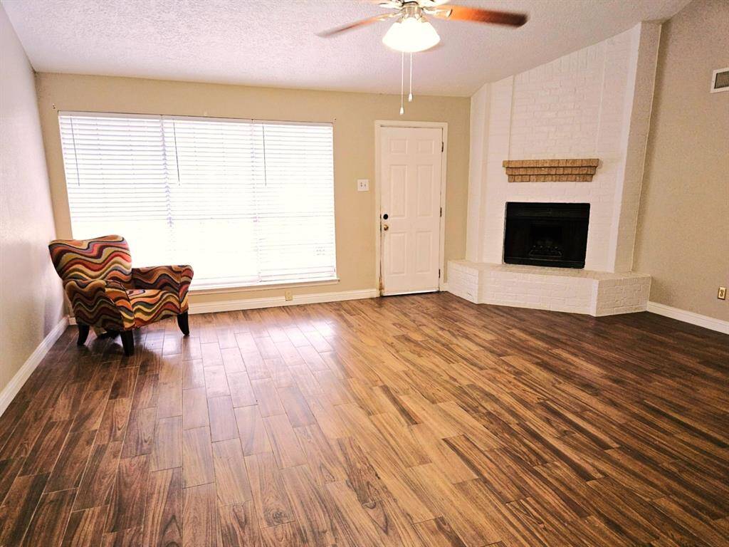 This inviting living room features warm wood look flooring, a cozy white brick fireplace, large windows for natural light, and a ceiling fan, offering a comfortable and stylish space.