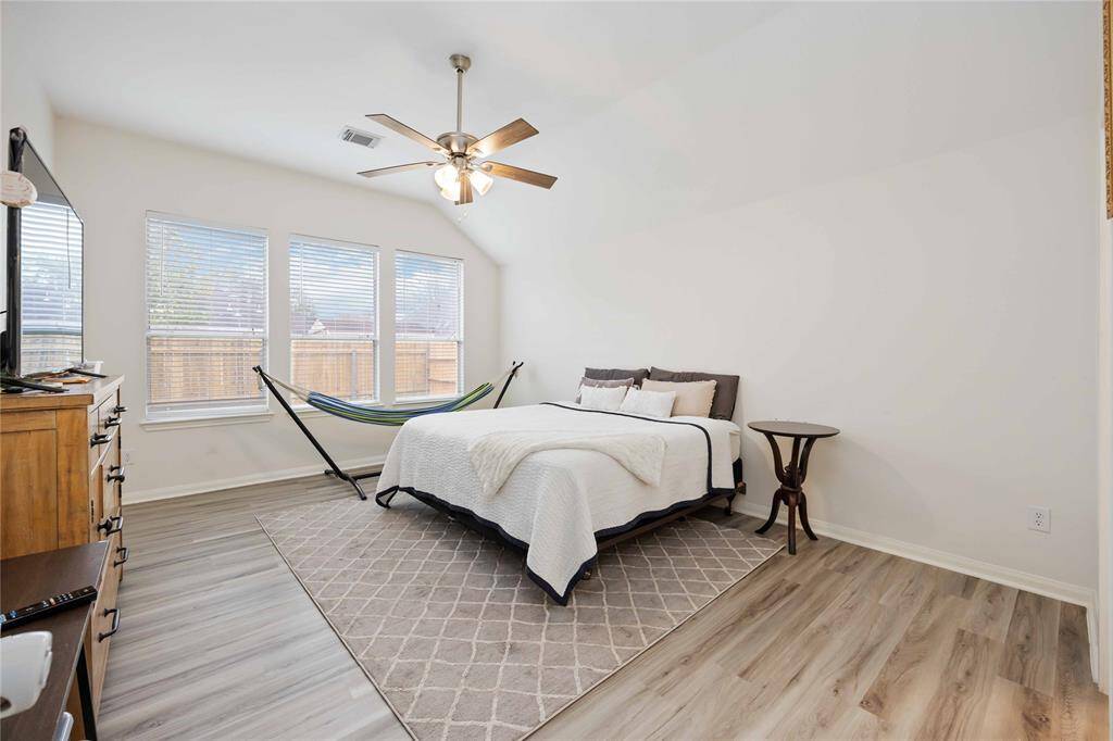 This bright primary bedroom features a modern ceiling fan, and large windows for natural light. The space is complemented by light wood flooring and a neutral color palette.
