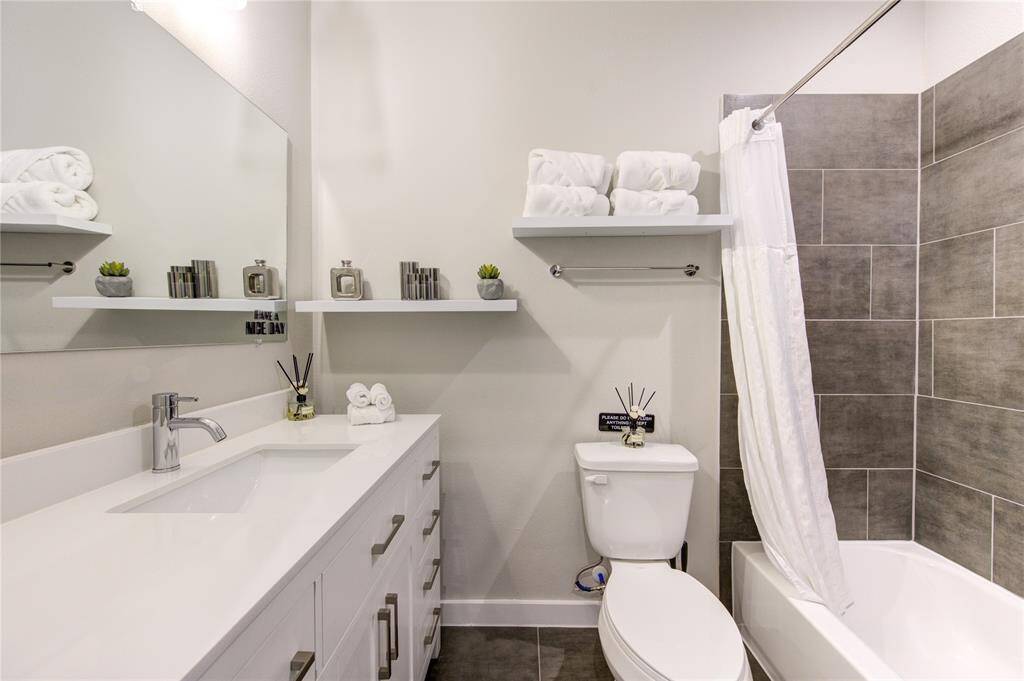 Modern bathroom in second floor with sleek white cabinetry and countertop. Features a large mirror, stylish gray tile in the shower, and open shelving for decor and essentials. Bright and inviting space.