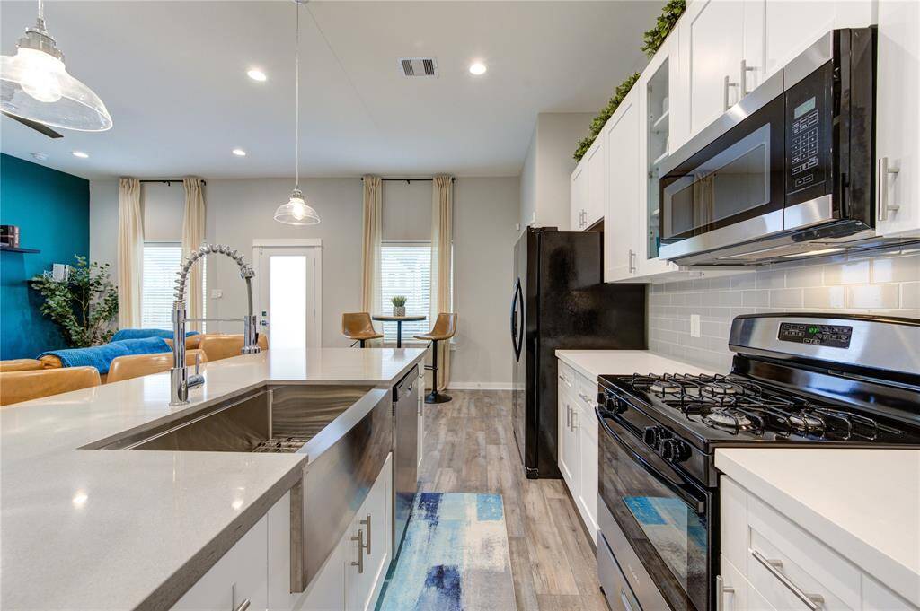 This modern kitchen features sleek stainless steel appliances, a spacious island with a large sink, and stylish pendant lighting. It opens to a bright living area with high ceilings, wood flooring, and a vibrant accent wall, creating a welcoming and functional space.
