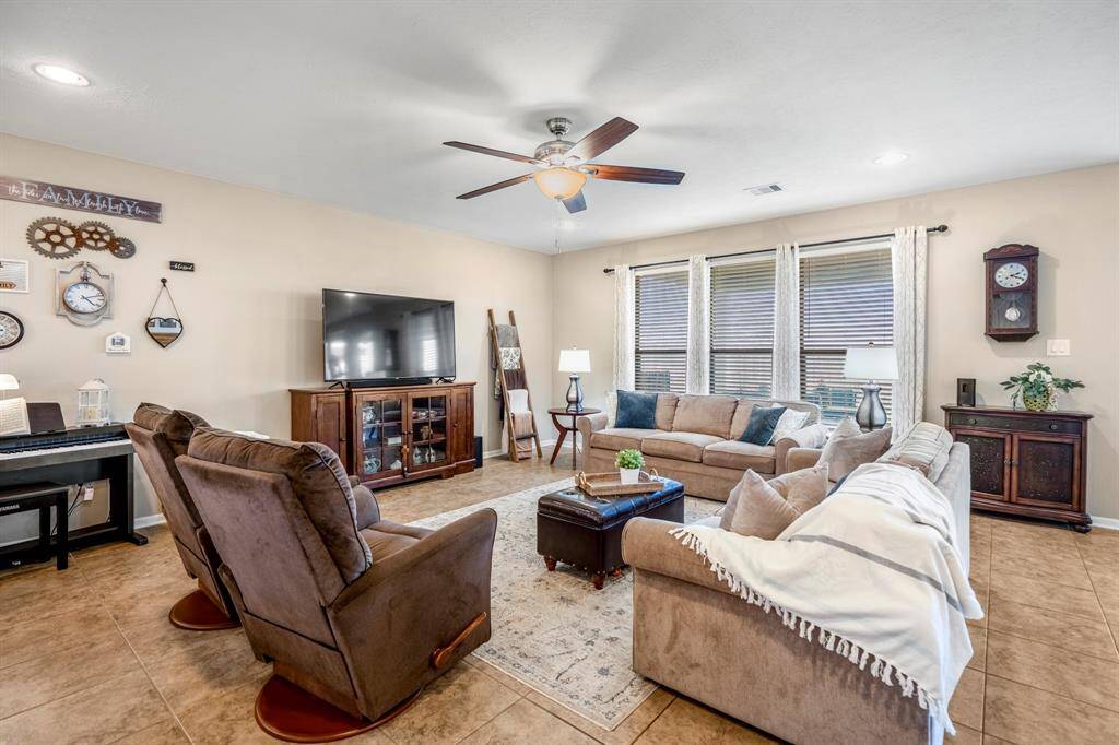 This spacious living room features large windows for natural light, a cozy seating area with plush sofas, a ceiling fan, and a large TV. The room is accented with elegant decor and a wall-mounted clock, creating a welcoming atmosphere.