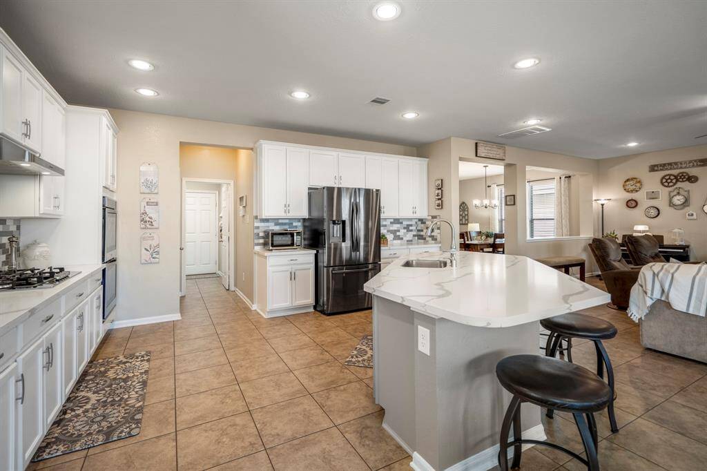 This spacious kitchen features modern white cabinetry, stainless steel appliances, and a large island with seating. It opens to a cozy living area, creating an inviting and functional space for cooking and entertaining.