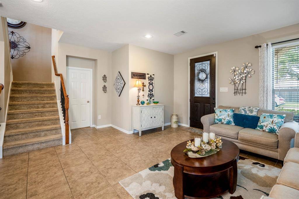 The space is well-lit with a window and a stylish lamp on a small side table. It has tile flooring and a welcoming staircase leading upstairs, with a warm, neutral color palette throughout.