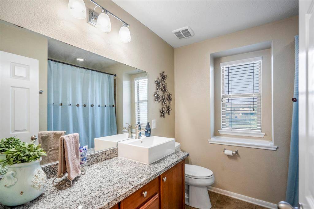 This bathroom features a modern design with a granite countertop, vessel sink, and wood cabinetry. It has a large mirror, stylish lighting, and a window for natural light.