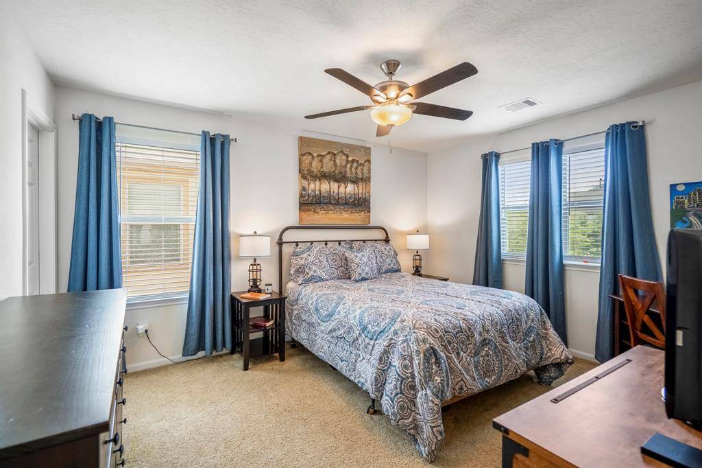 This bedroom is bright and inviting, featuring large windows, a ceiling fan, and neutral carpeting.