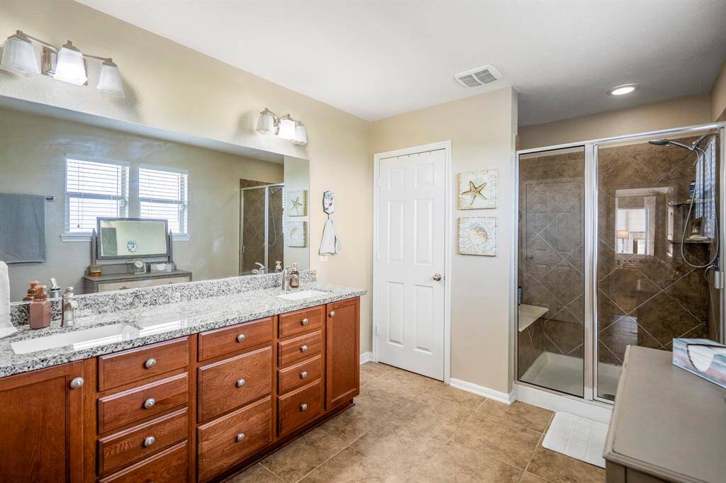 The primary bathroom features a spacious design with a double-sink vanity, granite countertops, and ample cabinet storage. It includes a modern glass-enclosed shower with tile accents and plenty of natural light from the windows.
