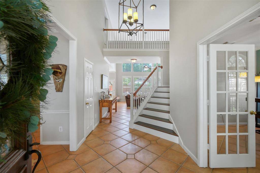 STEP INTO A GRAND FOYER FEATURING SOARING TWO-STORY CEILINGS, A WIDE STAIRCASE WITH ELEGANT WOOD ACCENTS, AND CLASSIC TERRACOTTA-STYLE TILE FLOORING.