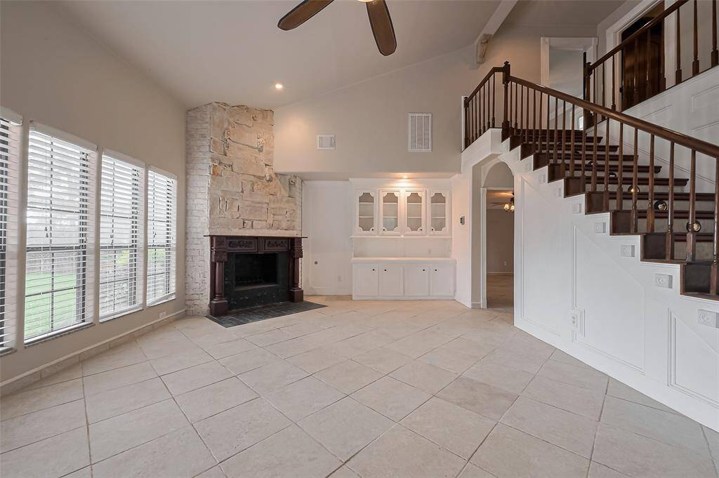This spacious living room features a dramatic stone fireplace with ornate wood mantel, high ceilings, and built-in cabinetry for added storage and display. A curved staircase with dark wood railings adds architectural interest and a grand focal point.