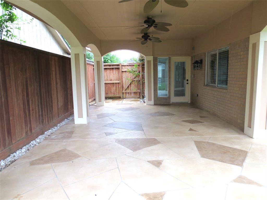 Spacious covered patio with a tiled floor, ceiling fans, and light fixtures. It's surrounded by a wooden privacy fence and brick wall, providing a cozy and private outdoor space.