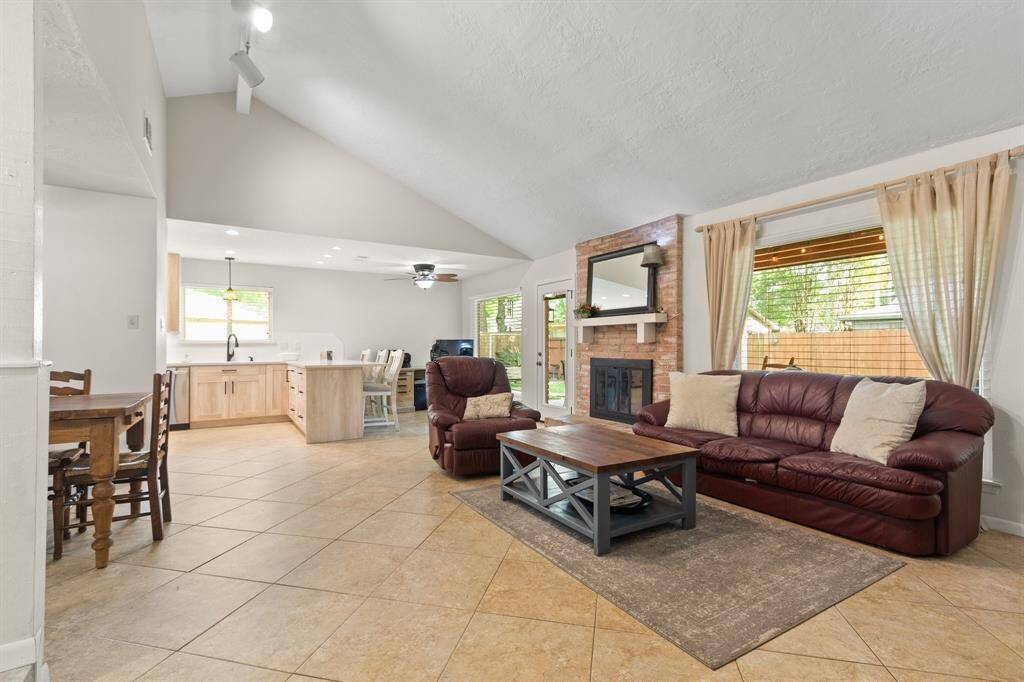 A spacious, open-concept living area with high vaulted ceilings, a cozy brick fireplace, and large picture windows providing natural light. The kitchen features modern cabinetry and countertop height eat in area, perfect for family gatherings.