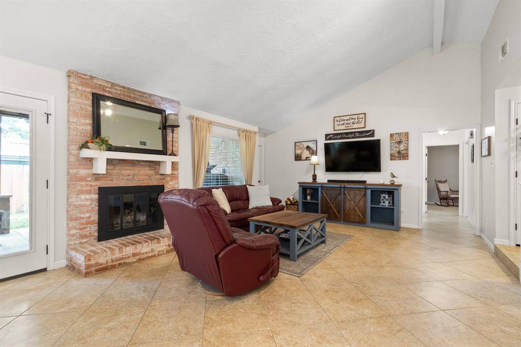 This spacious living room features a vaulted ceiling, a cozy brick fireplace, and large picture windows for natural light. The open layout includes tile flooring and a view into an adjacent room, enhancing the sense of space and flow.