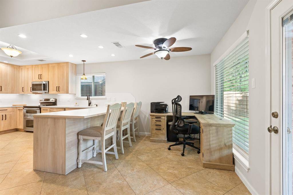 This bright and airy kitchen features modern light wood cabinetry, stainless steel appliances, and a spacious island with seating. Adjacent is a cozy nook with a desk, perfect for a home office, complemented by large picture windows and a ceiling fan.