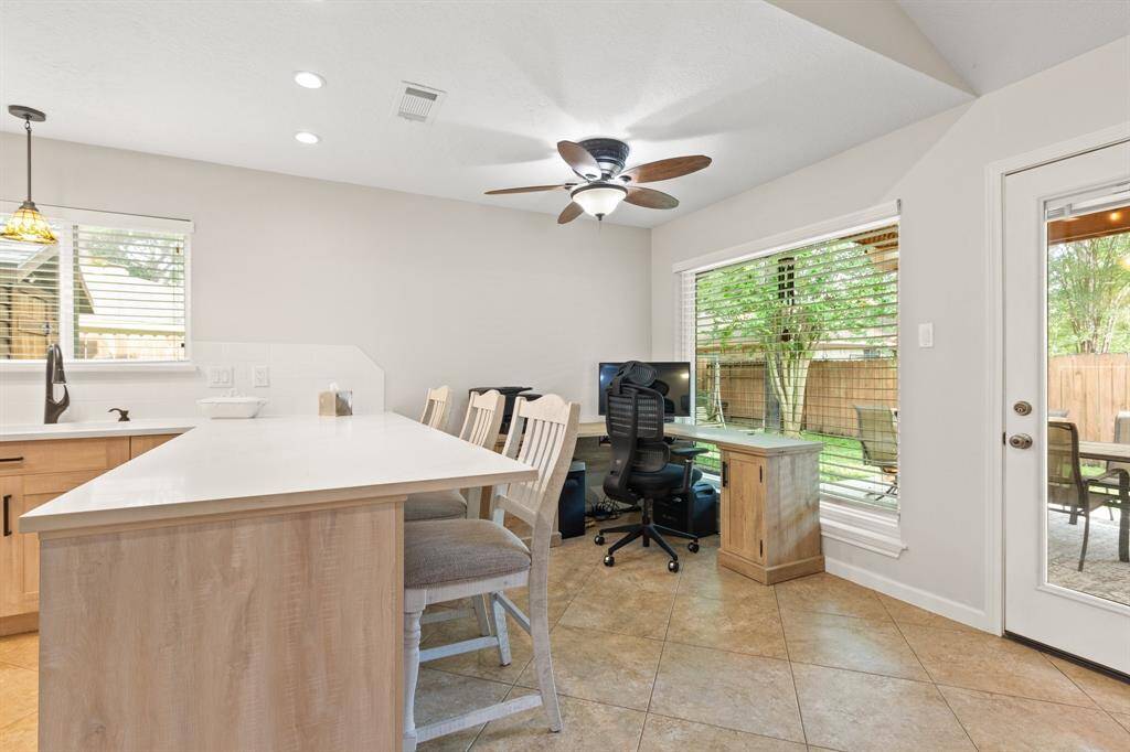 This bright and modern space features a kitchen with a large island and bar seating, an adjacent work area with wide picture windows offering views of the backyard. A ceiling fan and recessed lighting enhance the room's functionality and comfort.