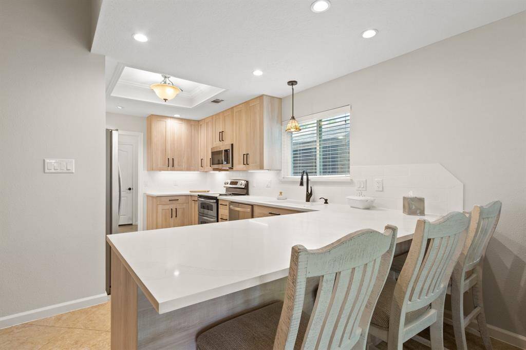 This kitchen features a modern design with light wood cabinetry, stainless steel appliances, and a spacious white with grey quartz countertop. The breakfast bar includes seating for three, and the room is well-lit with recessed and pendant lighting. A window above the sink provides natural light.